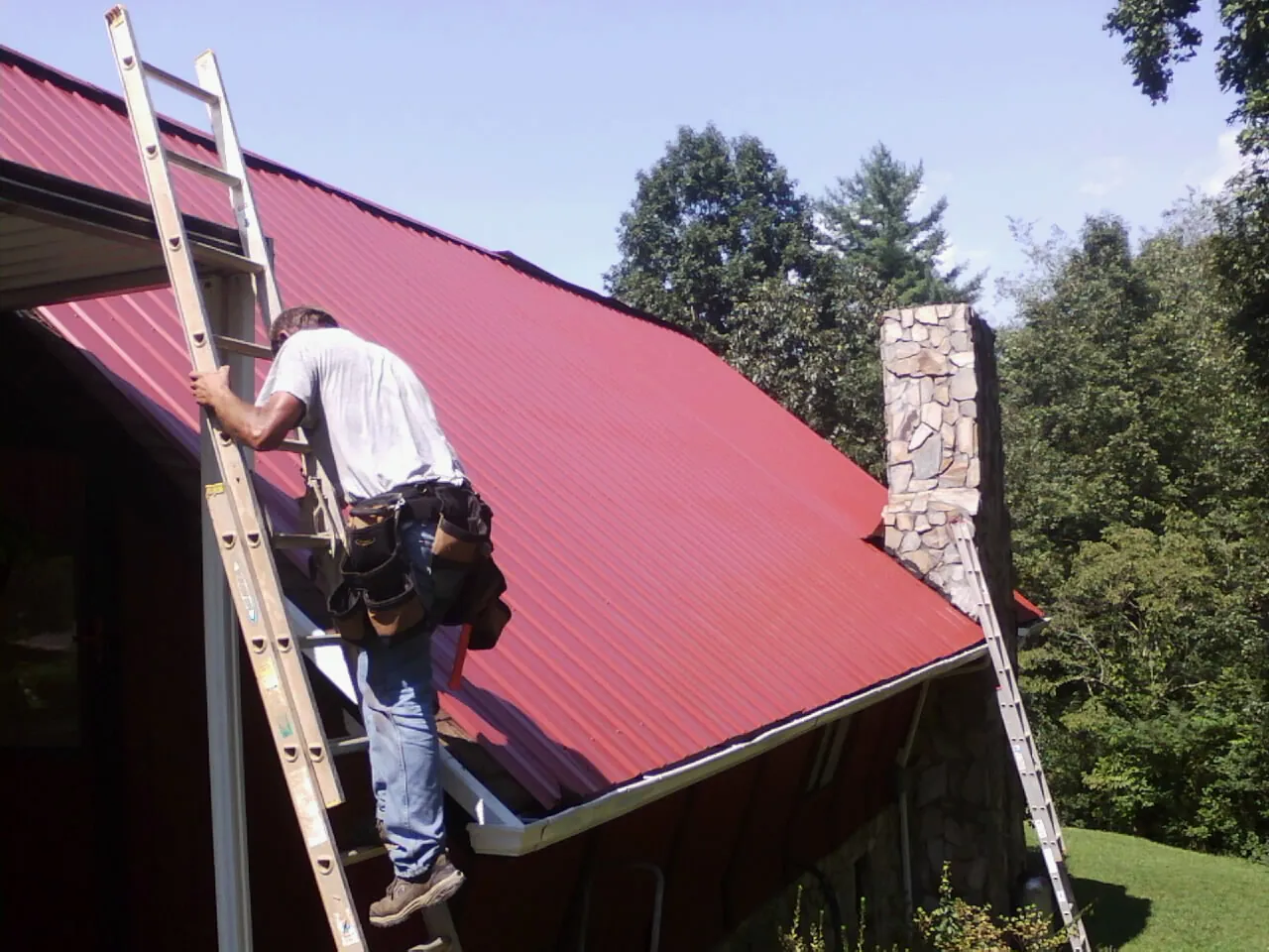 Roofer on a ladder beside a red metal roof and stone chimney — Soffit & Fascia Repair by Roofing Brothers in Gassaway